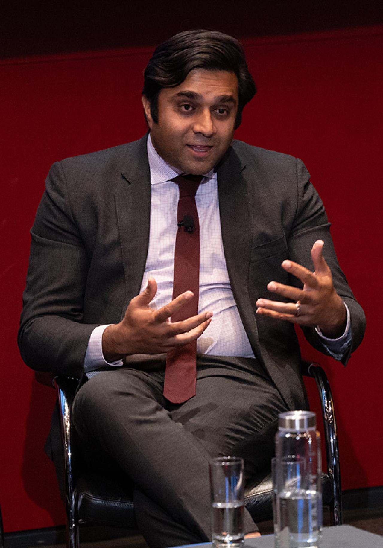 A seated man wearing a suit and tie gestures