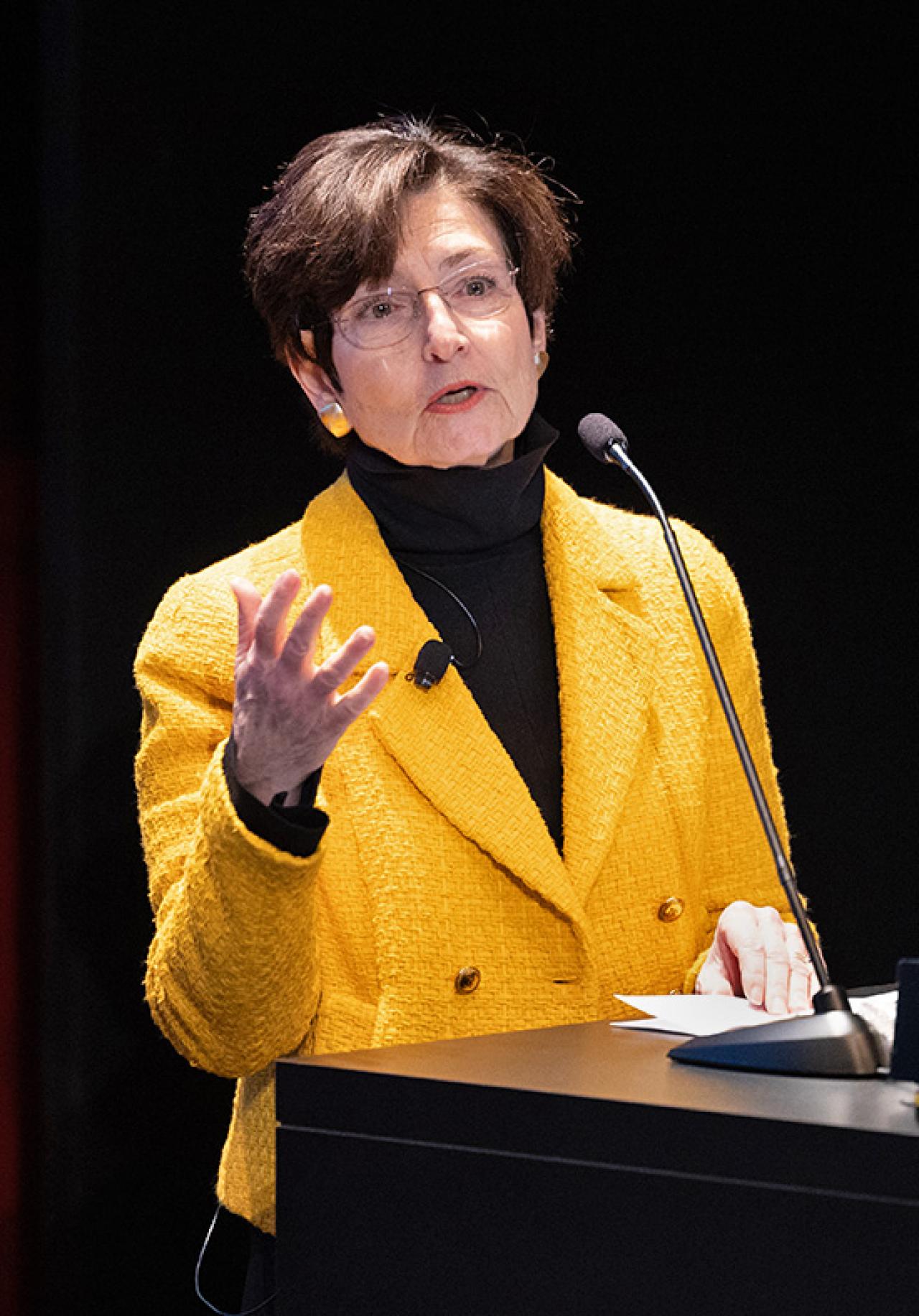 A woman in a yellow jacket speaks at a podium