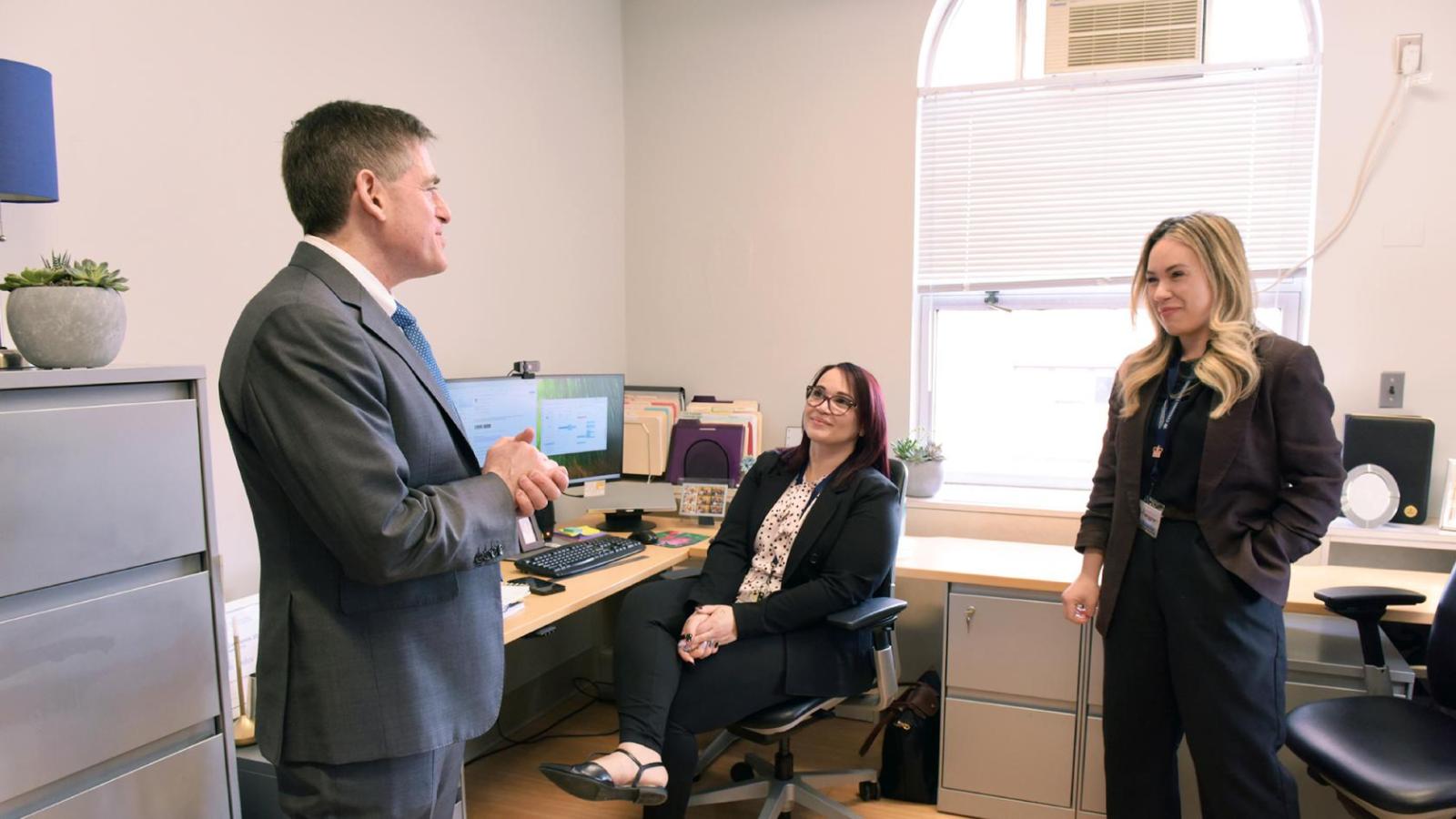 A man in a suit stands in an office occupied by two women, one seated and the other standing