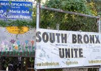 A chainlink fence with green leaves of a tree in the background. Signs read "Beautification This Site María Sola Community Greenspace" and "South Bronx Unite"