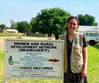 Natalie Andrasko beside a sign for her field practice site, Women and Rural Development Network in Uganda