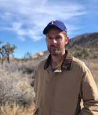A man dressed in a jacket and hat stands outside.