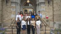 A group of 15 diverse, professionally dressed students stand and smile on the steps to the Allan Rosenfield Building