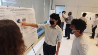 A student in a mask presents a scientific research poster to other students.