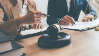 Two people sit at a table with a gavel on it. Only their torsos are visible. They have papers in front of them and pens in their hands.