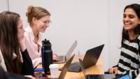 Three students sit around a table working on laptops and laugh