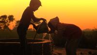 Two children at a well during sunset; one drinks from a bucket