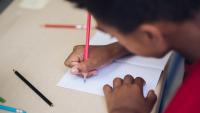 A boy seated at a desk and holding a pencil writes in a book