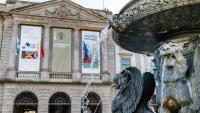 A working fountain with lions in the foreground in front of a university building in the classical style adorned with banners