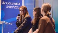 A woman wearing glasses speaks into a microphone as two other women look on. A sign in the background reads Columbia Mailman School of Public Health.