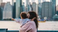 A woman holds a young child who points across water at at the Manhattan skyline