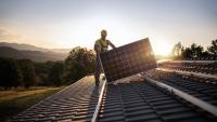 A worker installs solar panels with the sun and mountains behind him