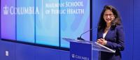 A woman wearing blue speaks at a podium in front of a blue background with the words Columbia Mailman School of Public Health visible