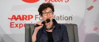 A seated woman speaks into a microphone with the logo AARP Foundation Experience Corps - 30 Years behind her