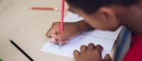 A boy seated at a desk and holding a pencil writes in a book