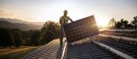 A worker installs solar panels with the sun and mountains behind him